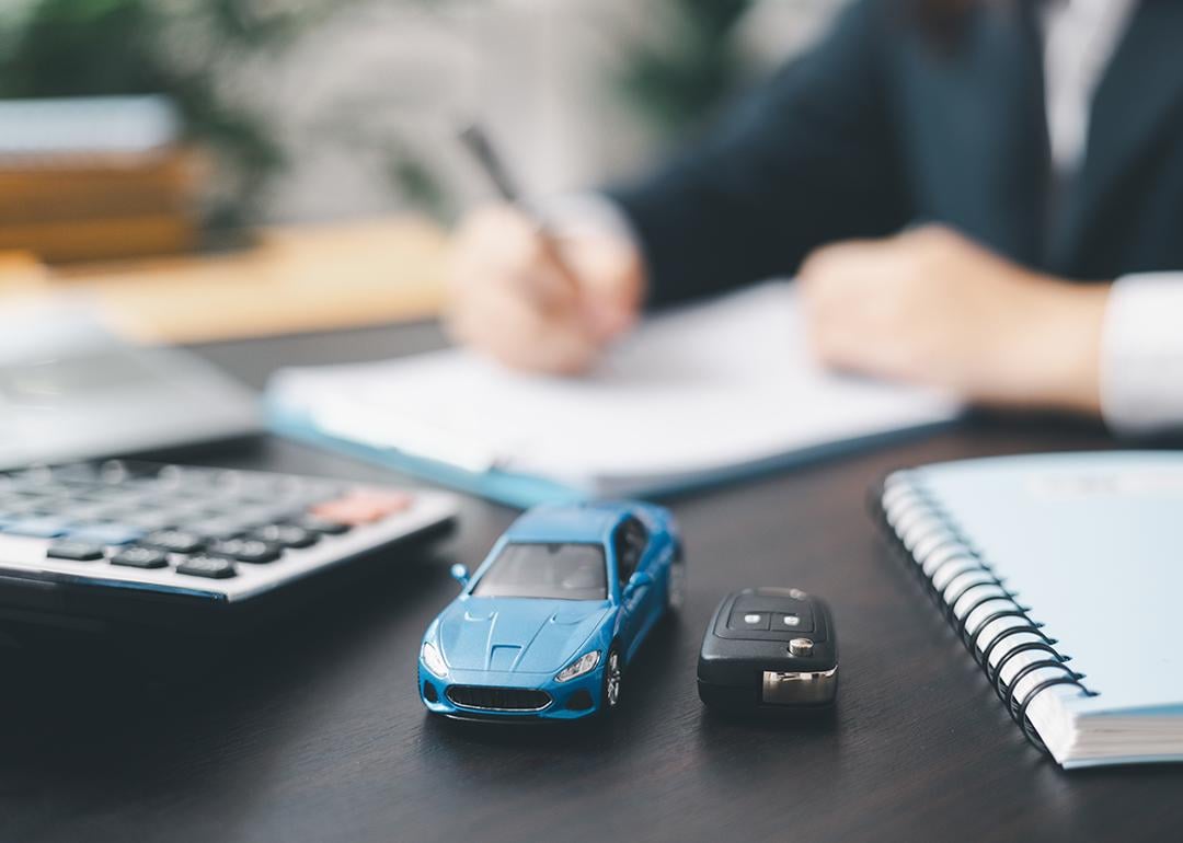 A small, blue car figure beside a car key in front of a car agent reviewing contracts.