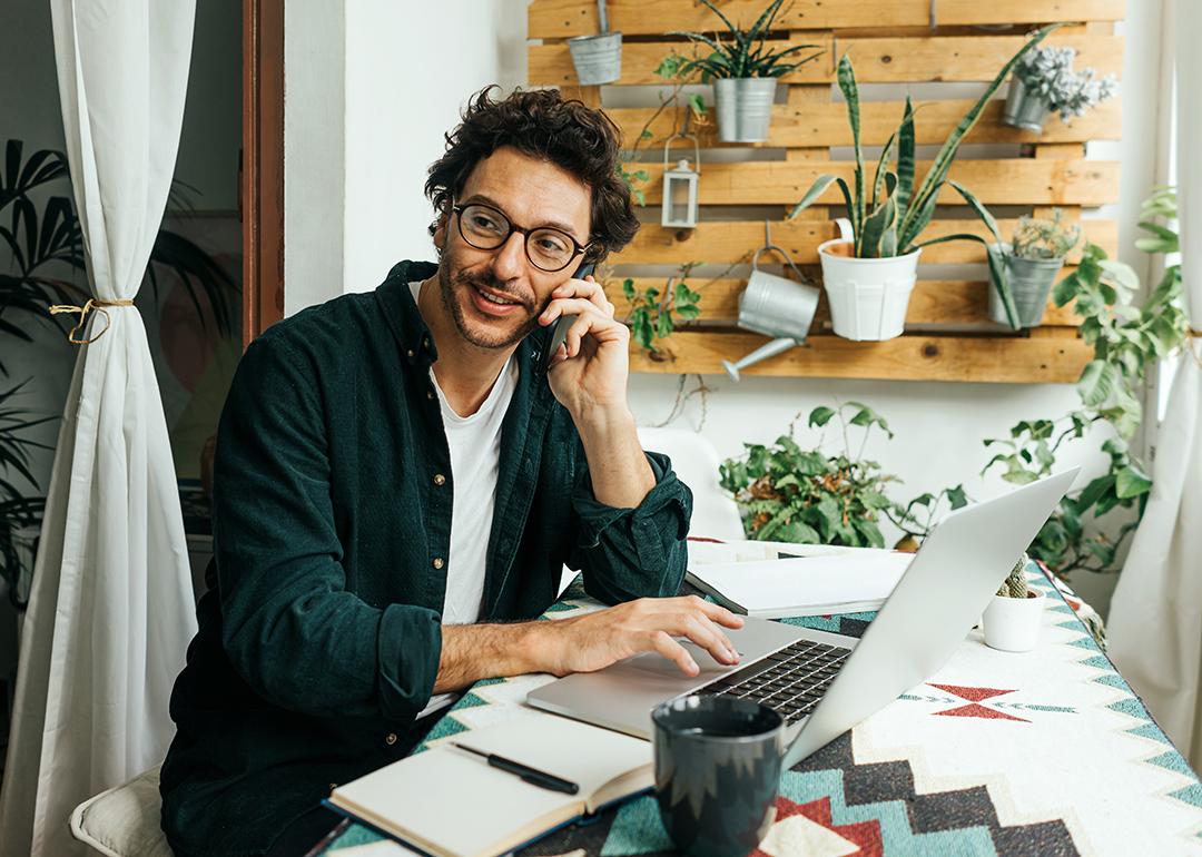 A young man on phone call and using laptop to work remotely from home.