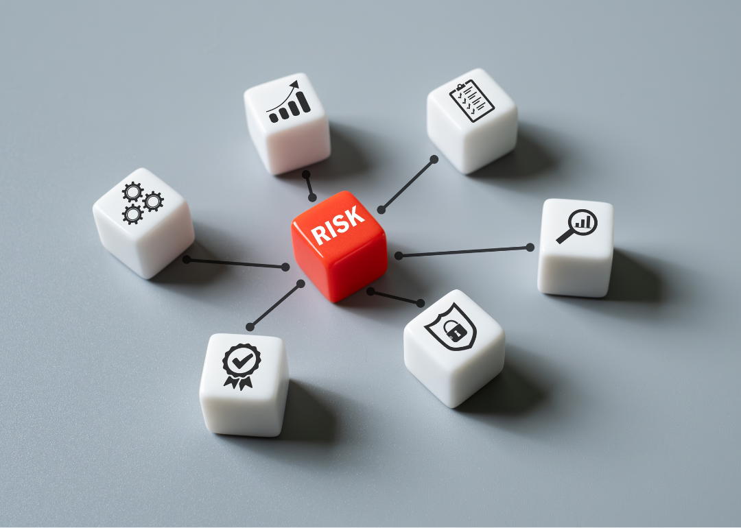 One red die with the word risk on it, positioned in in the middle of six white dice with symbols, including three cogs, an upward bar graph, a checklist, a magnifying glass over a rising bar graph, a security shield with a lock in the middle, and a ribbon with a check mark.