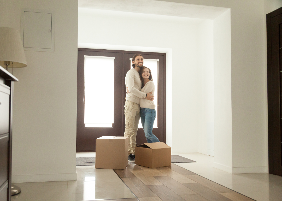 A hugging couple stand in the house in front of the door with two cardboard boxes in front of them.