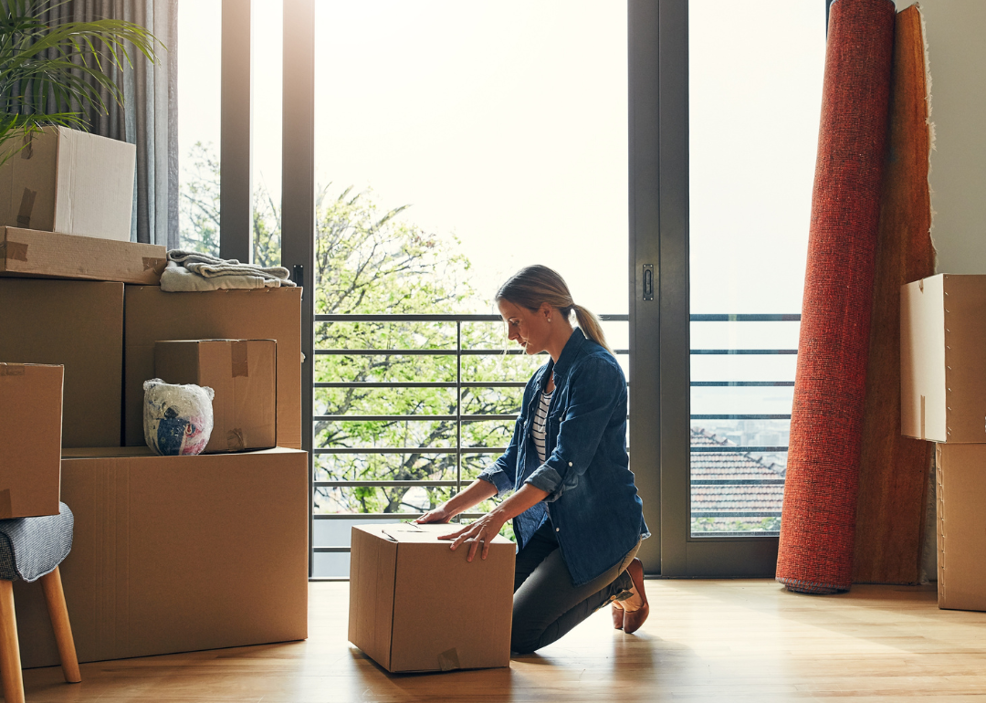 A women kneels in front of a cardboard box in a bright room with other boxes and household items like lamps and furniture. 