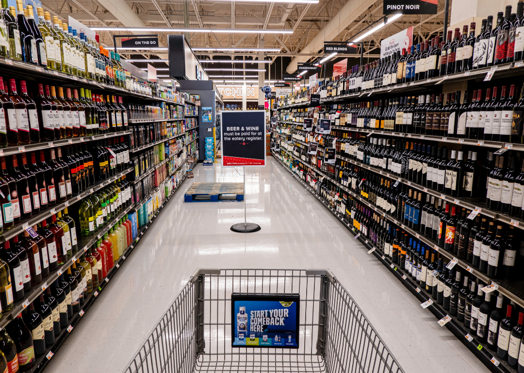 A shopping cart in a grocery store aisle filled with wine bottles.