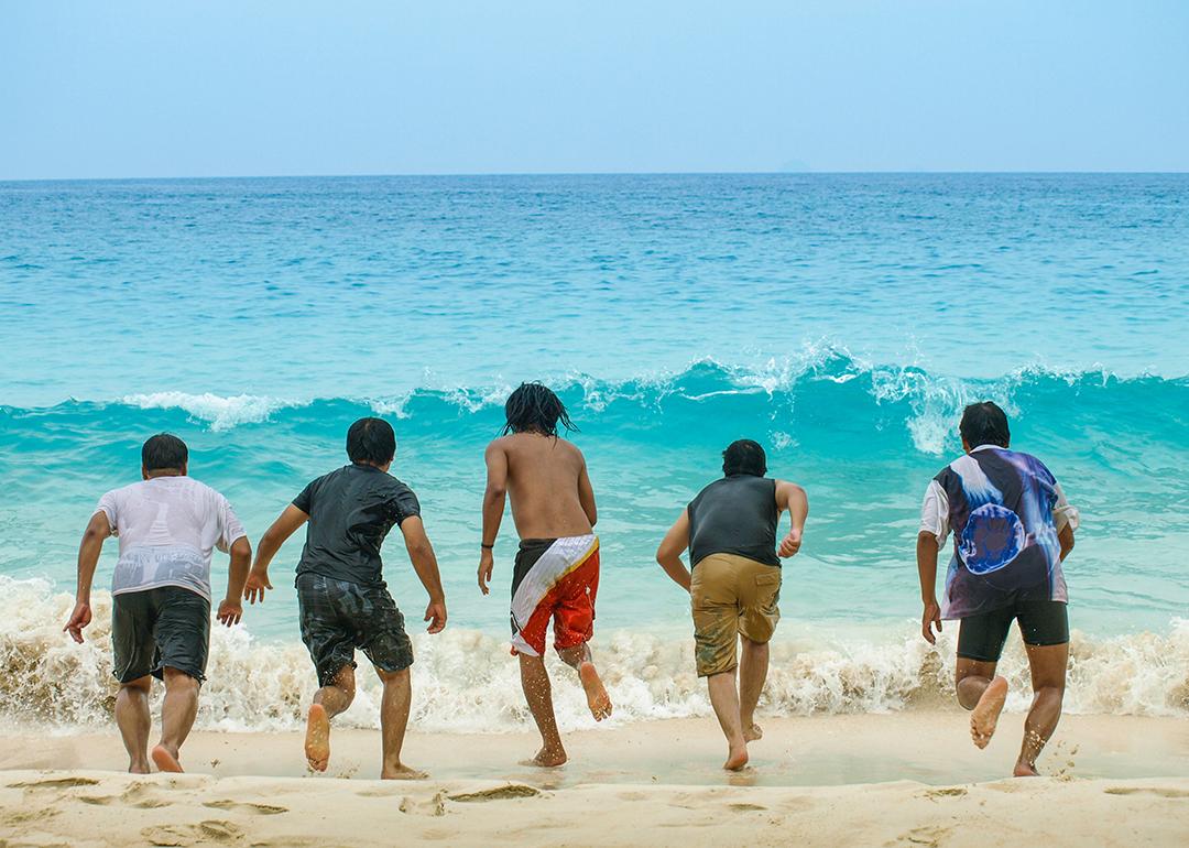 A group of five friends lined up to run towards a sea wave from the beach.