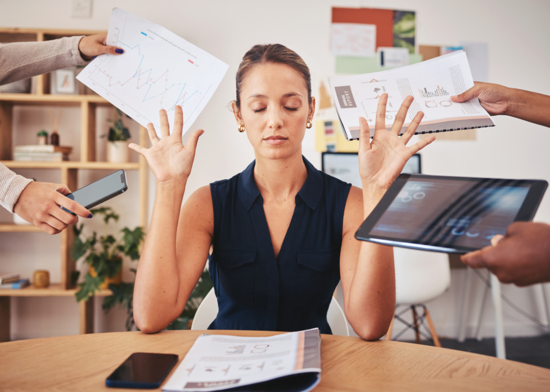 A women sitting in an office closes her eyes and puts her hands up as people hand her things including a phone, a computer, a paper with a graph on it, and a project notebook.