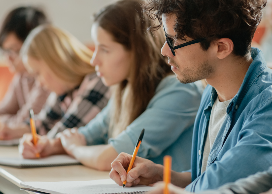 Teenage or young adult students seated in a row, taking notes. 