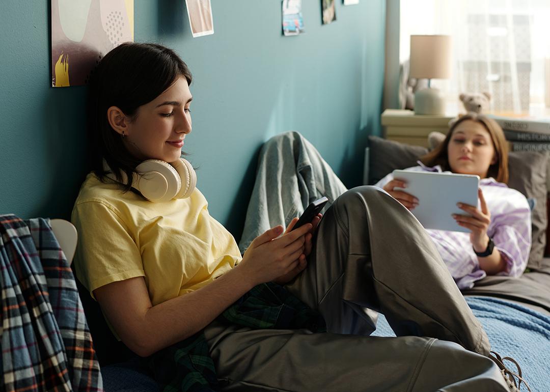 Two teenage girls relaxing on a bed and using smart devices to browse the internet.