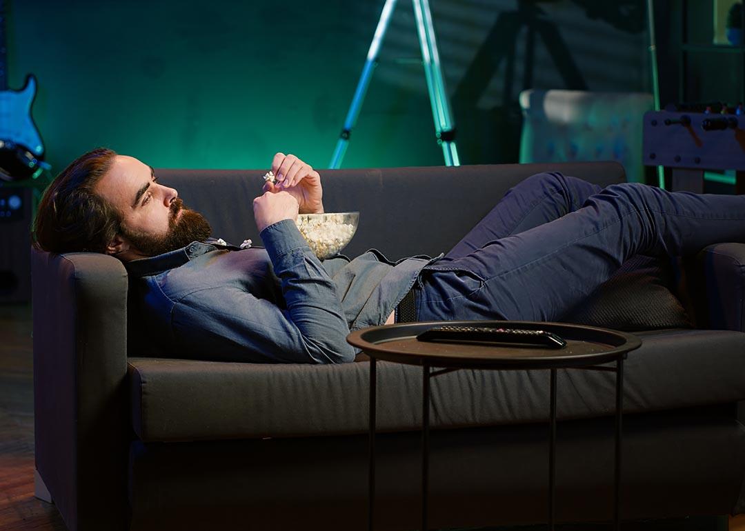 A bored man lying on couch and eating popcorn.