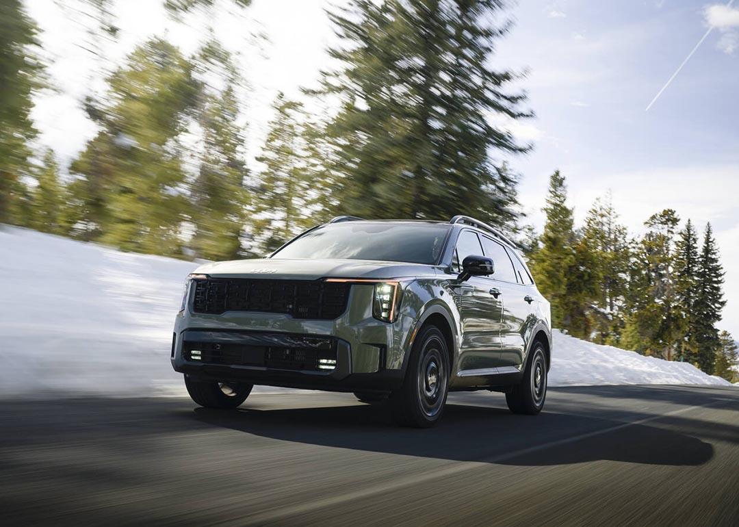 Low-angle view of earth-toned green 2026 Kita Sorento driving on mountain road with motion-blurred snow and pine trees in the background.