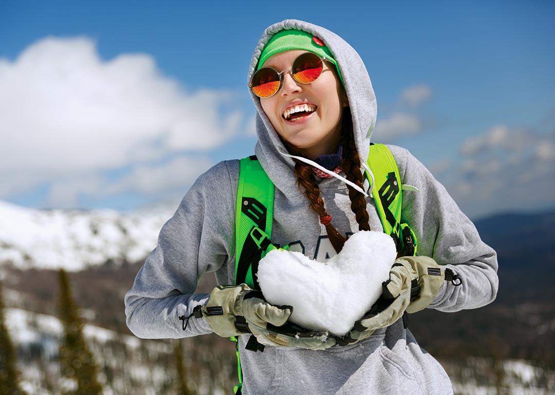 A young woman in snowboarding gear holding a heart shape made of snow.