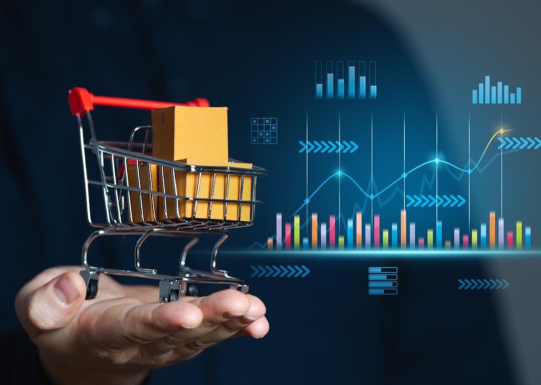 Concept of retail sales illustrated by perosn holding miniature shopping cart with packages inside; chart with growth figures and data in the background 