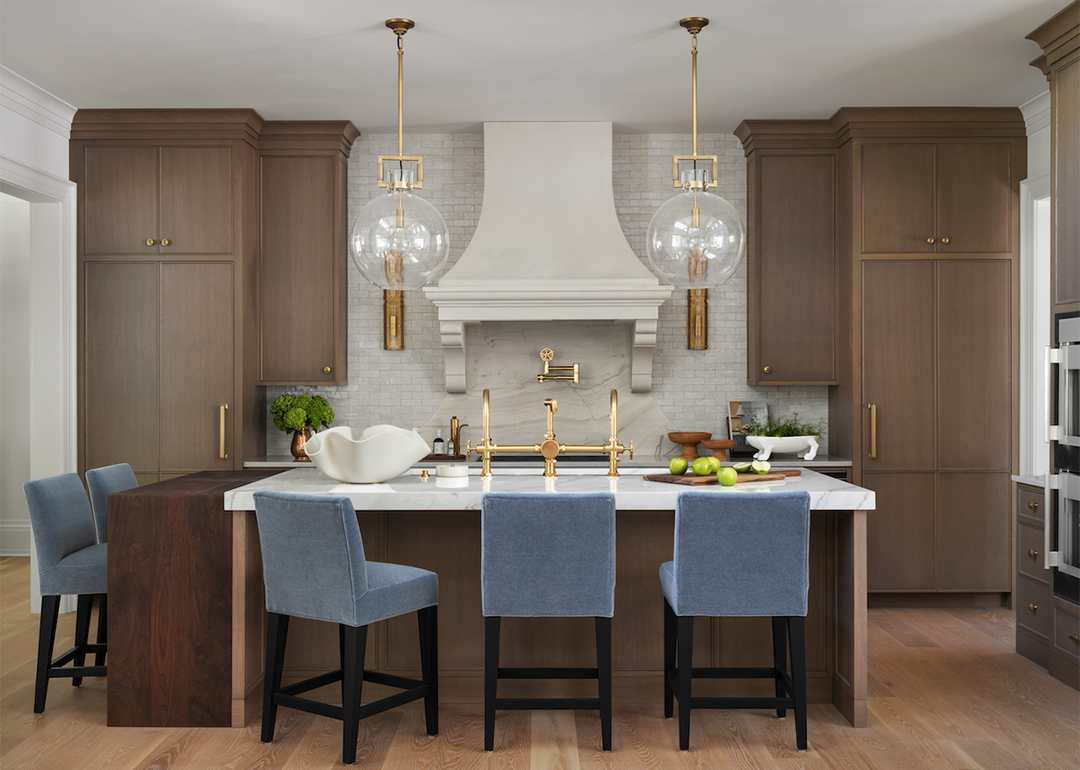 A kitchen with wooden cabinets, modern gold fixtures and light blue velvet chairs around a center island.