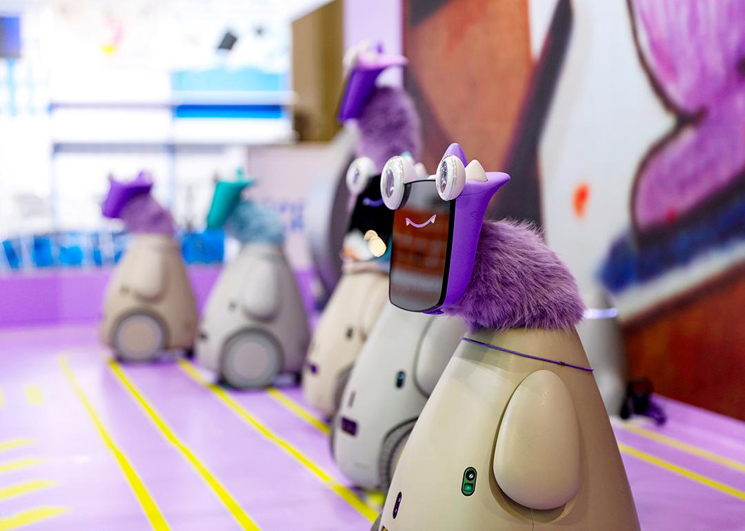 A row of 5 "robot pets" with playful googly eyes affixed on tablet faces and bright-colored faux fur necks on display at a convention. 
