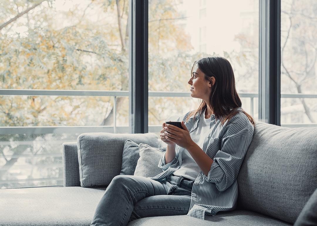 Young woman sitting on a sofa and having a cup of coffee while looking out the window at home.