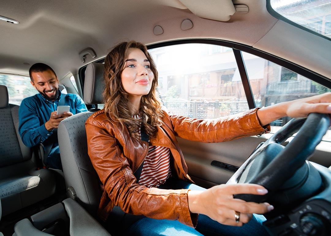 Wide shot of long-haired driver in brown leather jacket in the driver's seat with a passenger in backseat using phone.