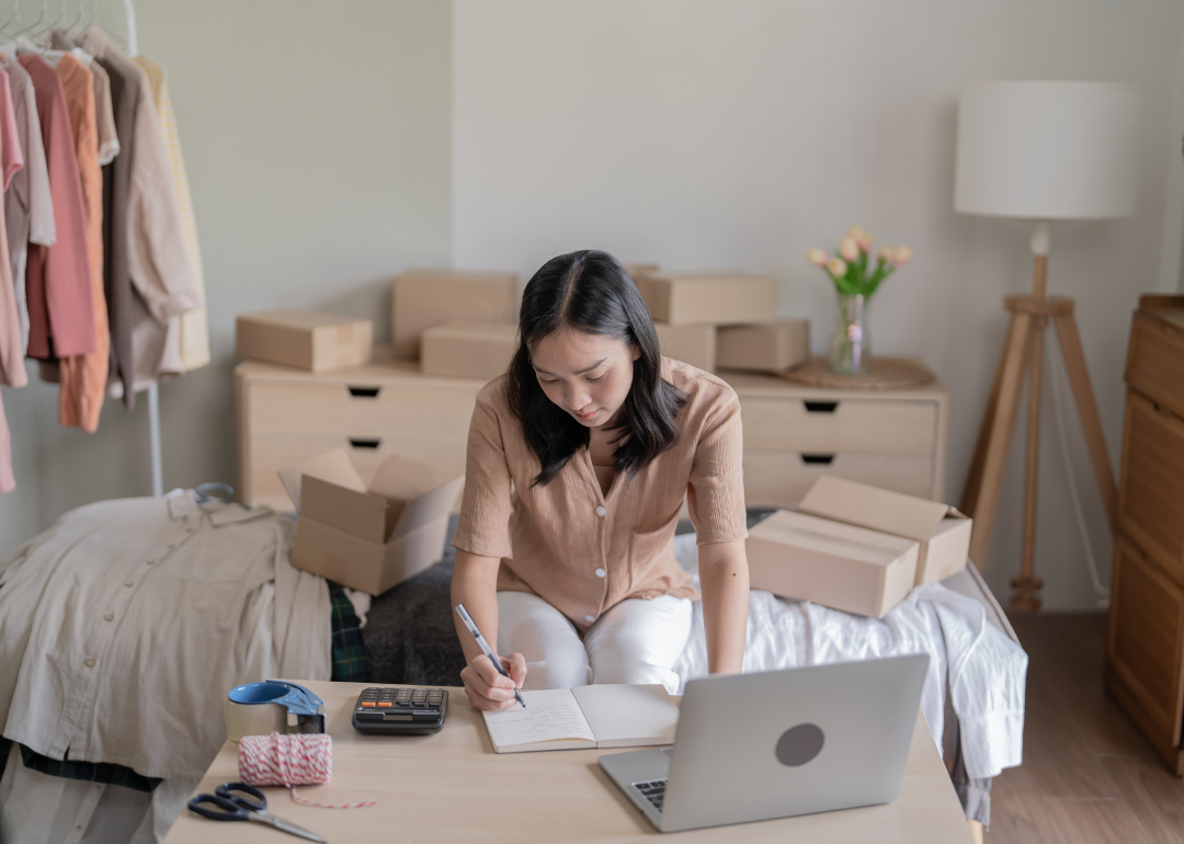 A woman writing in a notebook on a desk with a calculator, a laptop, scissors, tape and string, in a room with clothes hanging on a rack and cardboard boxes stacked on various surfaces.