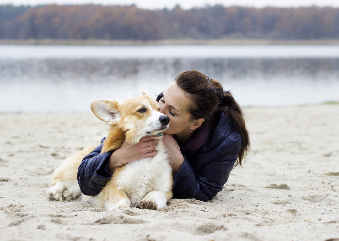 A woman lays on the sand at a beach near a lake, hugging and kissing a corgi laying next to her.