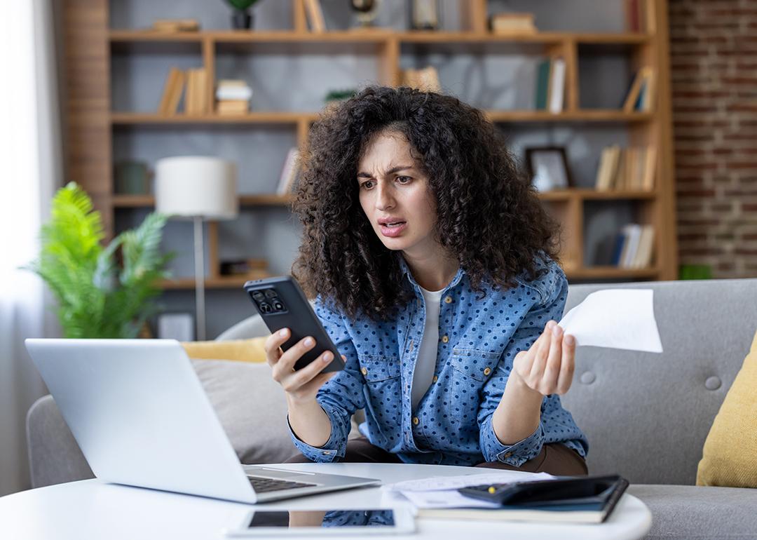 A young woman at home stressfully looking at her smartphone while calculating expenses.