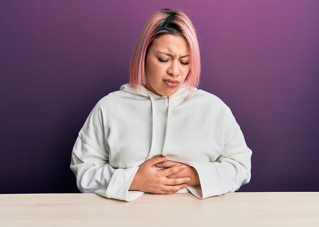 A woman with pink hair sitting with her hands on her stomach, feeling unwell.