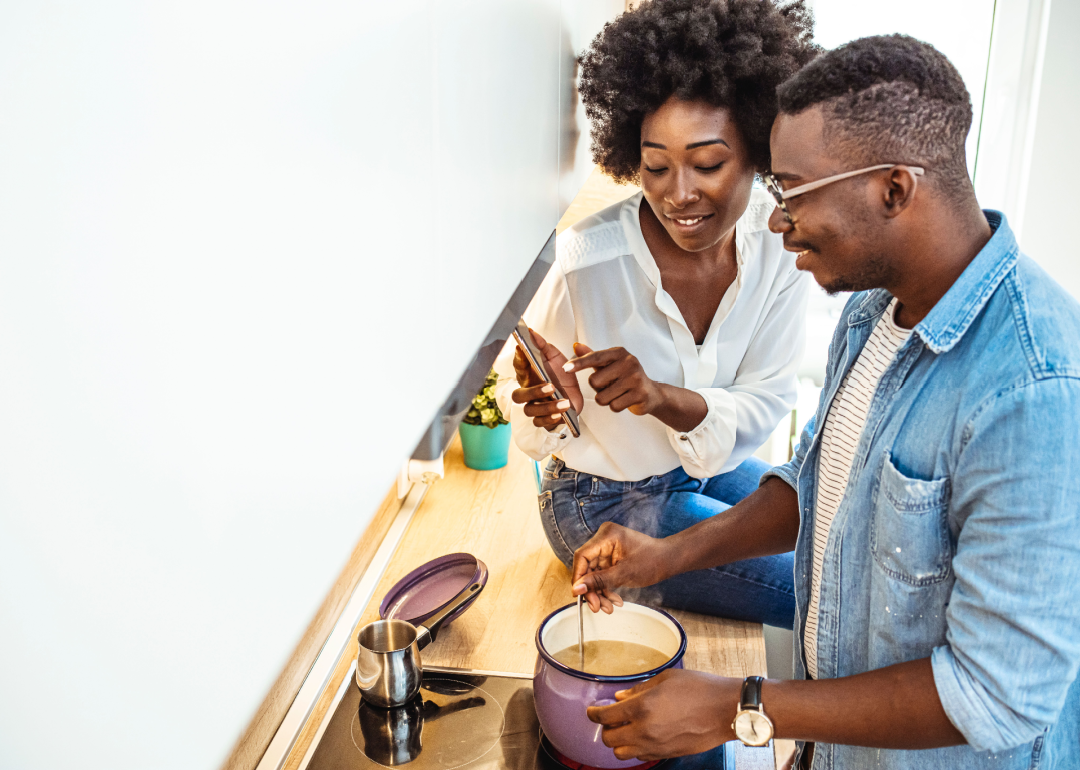 Young couple is using a smartphone and smiling while cooking in kitchen at home.