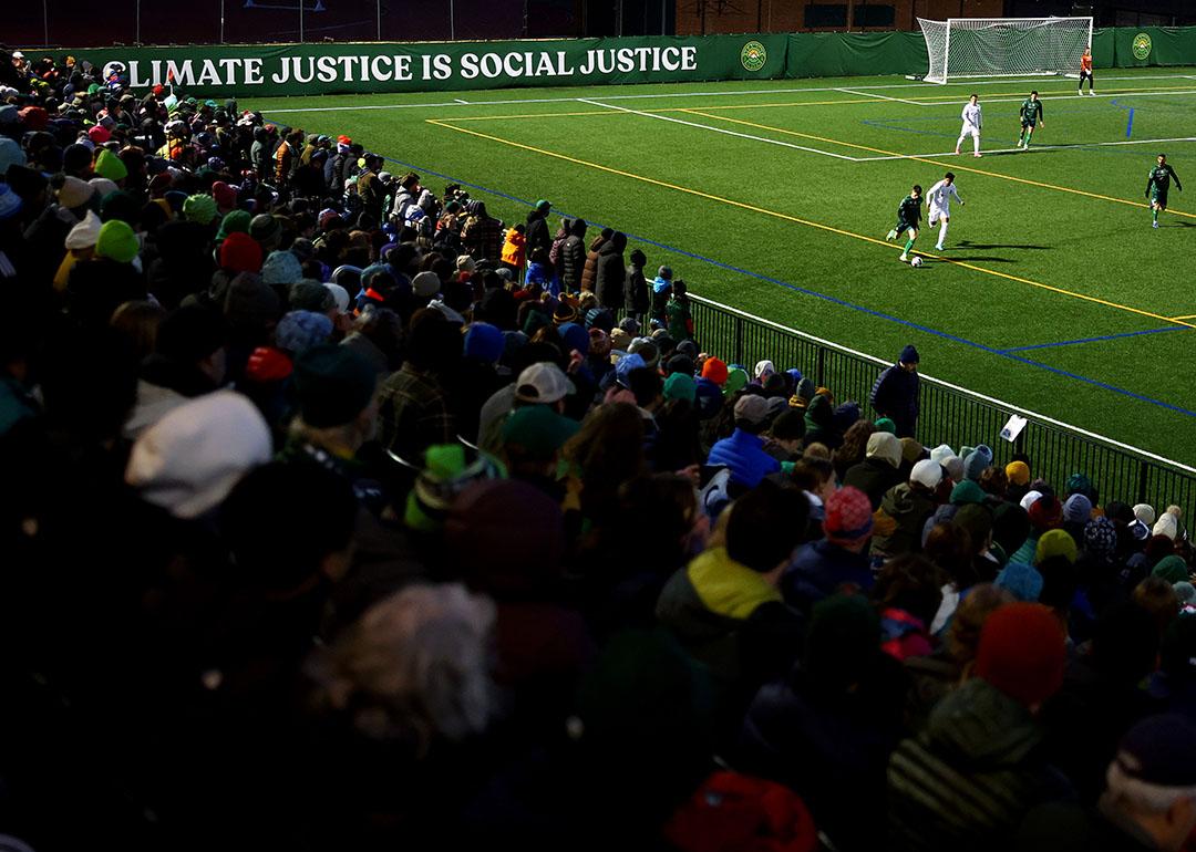 A view from the stands of the match with fans visible in the shadows on the bottom left and the green soccer pitch on the top right, with the words "Climate justice is social justice," displayed behind the goal during the  between the Vermont Green FC and Lexington SC during the 2024 U.S. Open Cup at Virtue Field on March 19, 2024 in Burlington, Vermont. 