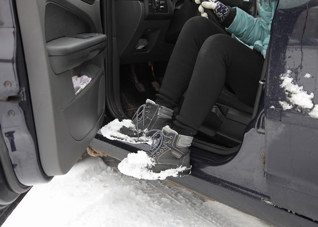 A young woman shaking snow off her boots while getting out of a car.