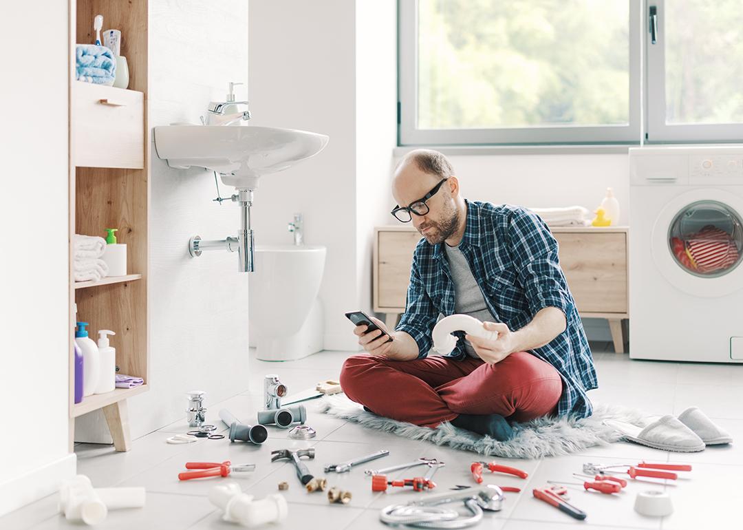 A man watching something on his smartphone while repairing a sink at home.