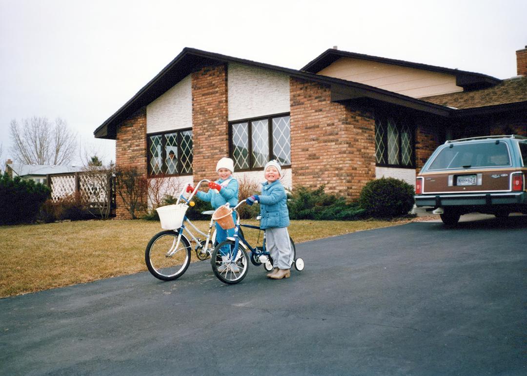 A vintage photo of two 80s siblings, boy and girl, riding bikes in their driveway.