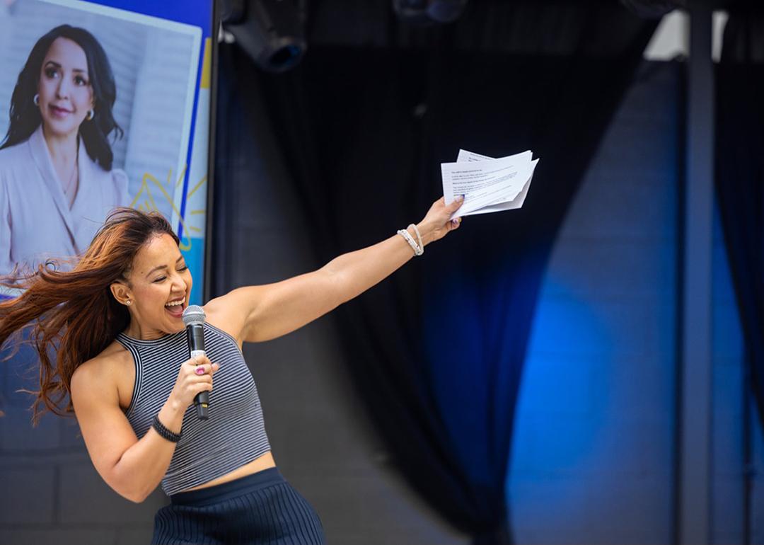 Julia Collins who is the founder and CEO of Planet FWD, speaks onstage with one hand holding a microphone and the other arm raised, her portrait is visible on a screen in the background.