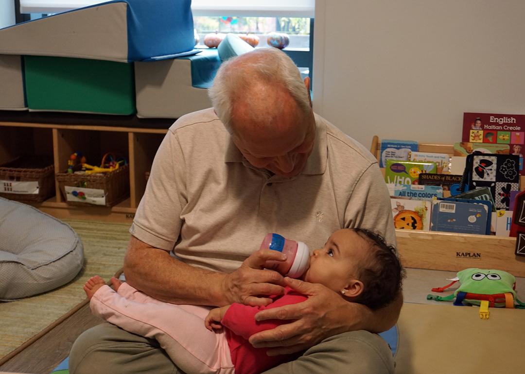 Teacher Herb Hickey holds a baby while she drinks from a bottle.