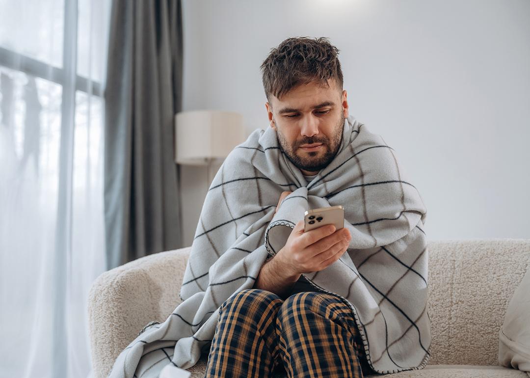 A sick man at home wrapped in blanket and looking up information on his phone.