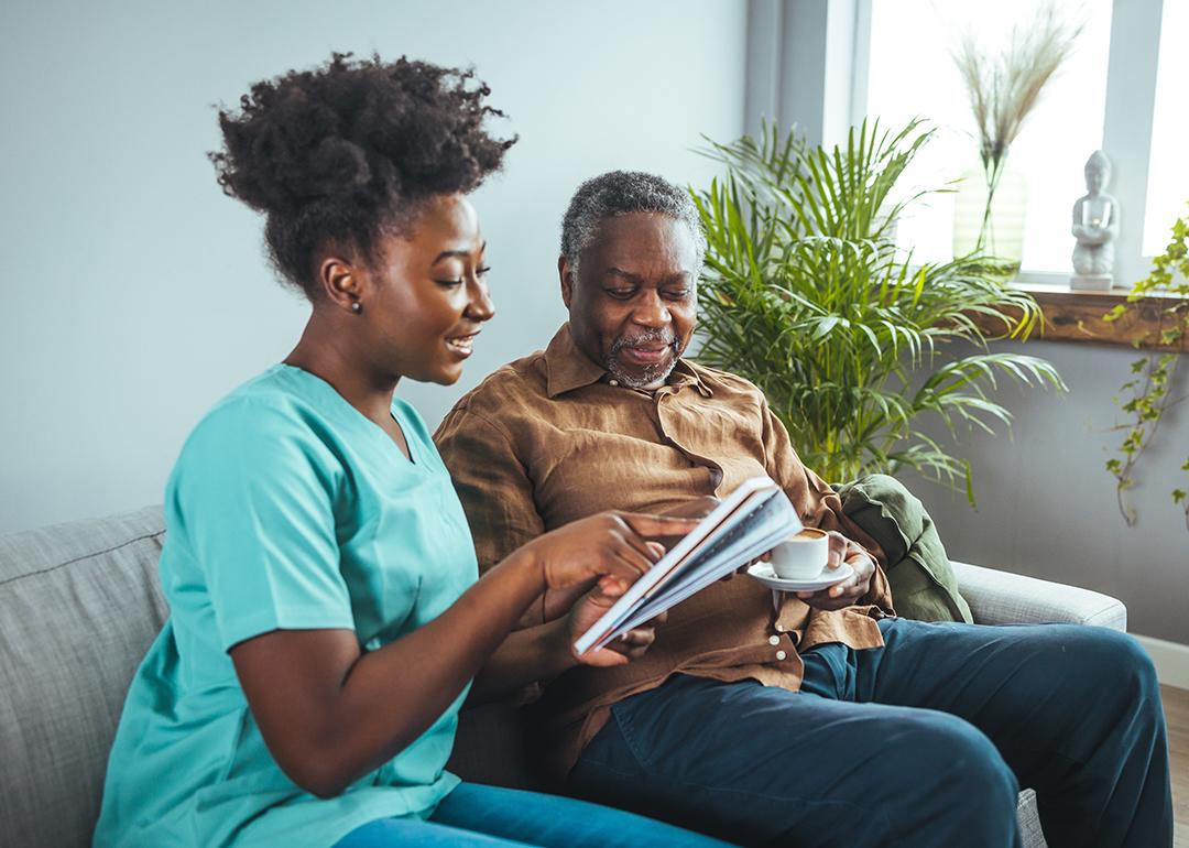 A black mid-adult nurse attentively aids an elderly black male with his reading material.