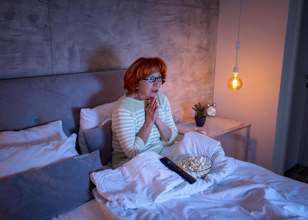 A senior woman watching a scary movie on the TV from bed.