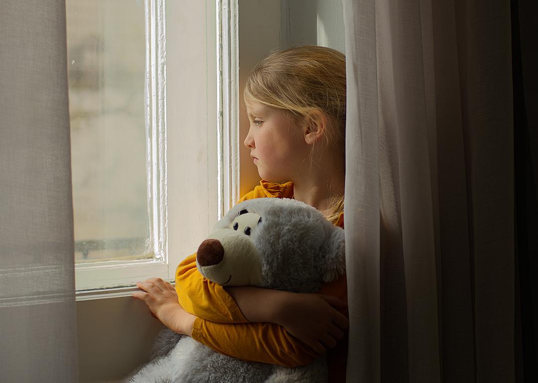 A kid with a stuff toy looking out in a window. 
