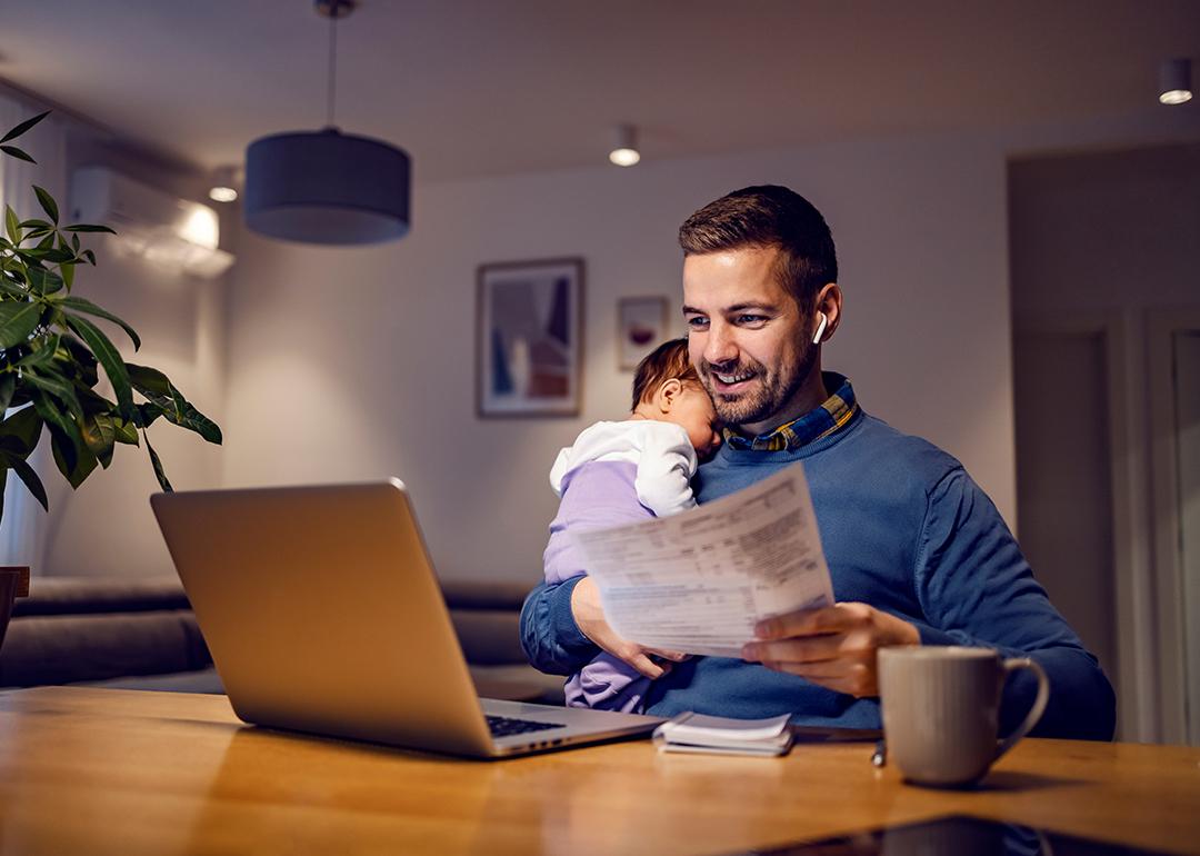A young father carrying his baby asleep while working remotely from home.