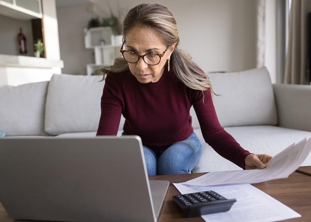 A focused senior woman holding financial documents and browsing information on a laptop.