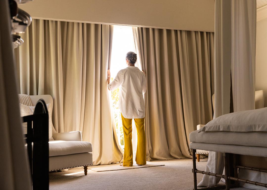 A senior woman standing by the large window of a cozy living room.