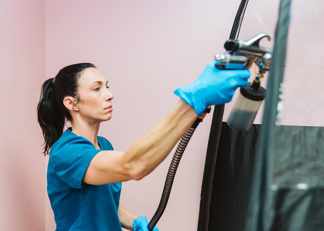 A beauty technician applying spray tan to a client inside a black booth.