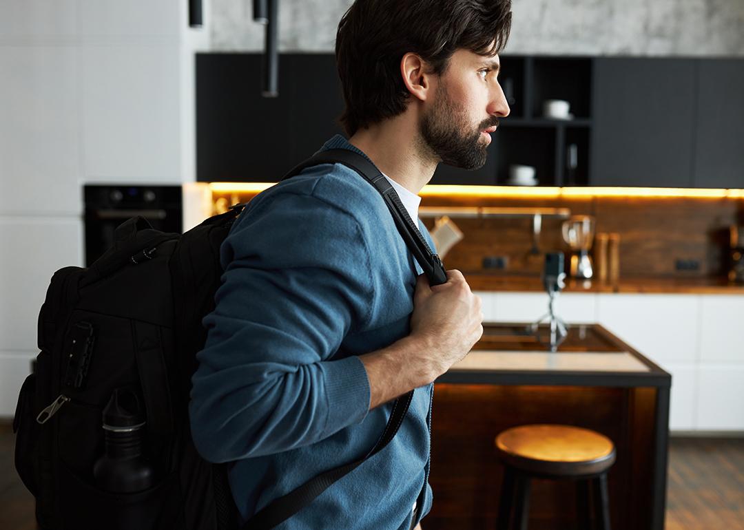 Man at home in a blue cardigan and putting on a black backpack preparing for a trip.