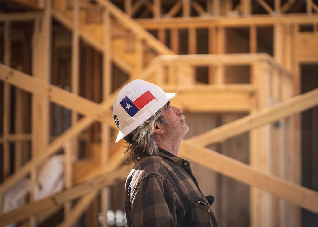 A construction worker wearing a hard hat with the Texas flag and looking up inside a wooden house construction site.