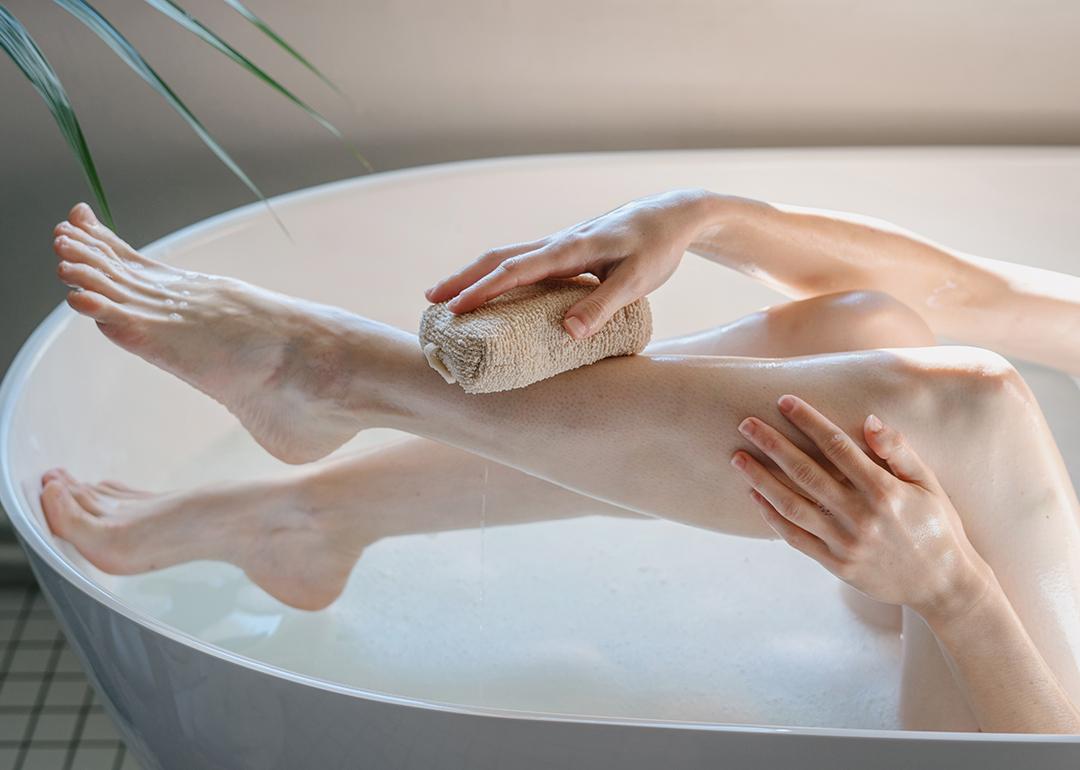 A cropped view of a person washing her legs in a bath tub with a body sponge.