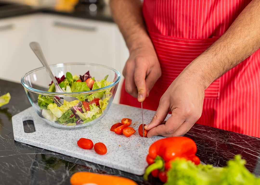 A person slicing cherry tomatoes to a greens salad in the kitchen.