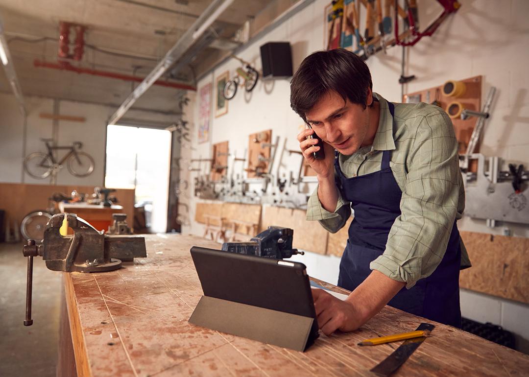 A male bicycle small business owner on the phone while reviewing information on a laptop in a workshop.