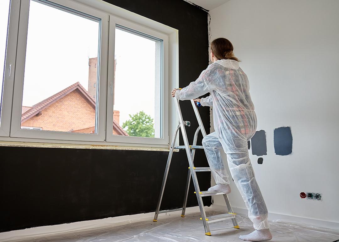 Woman in protective wear climbs a ladder to repaint a wall.