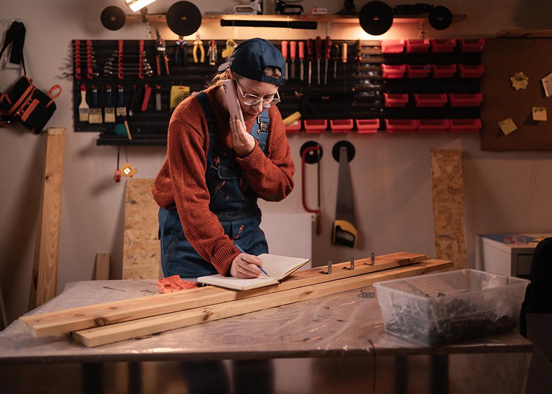 A female carpenter writing down notes while talking on the phone in a workshop.