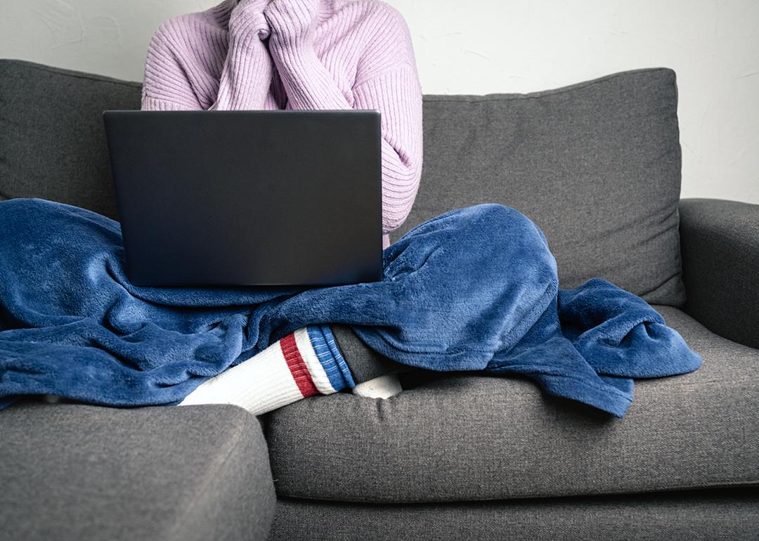 Person wearing a knitted sweater and socks with a blanket on while working and sitting on a sofa.