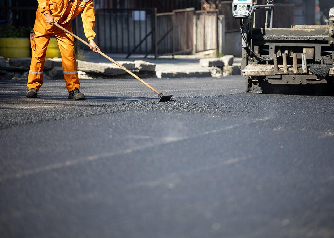 A construction worker in an orange uniform spreads freshly poured asphalt on a road surface.