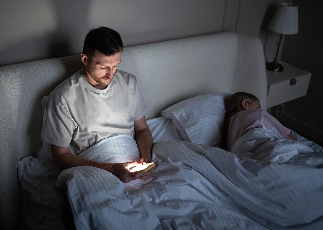 A man awake late at night and scrolling through his phone beside his asleep wife.