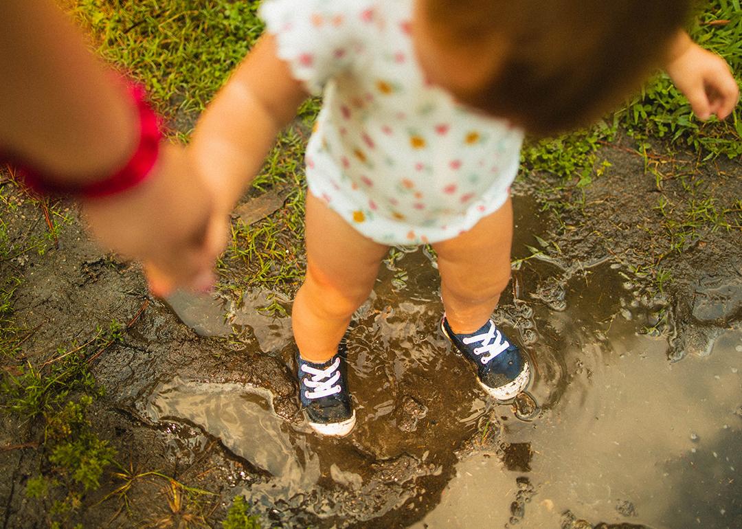 A toddler holding mother's hand while playing in a mud puddle.