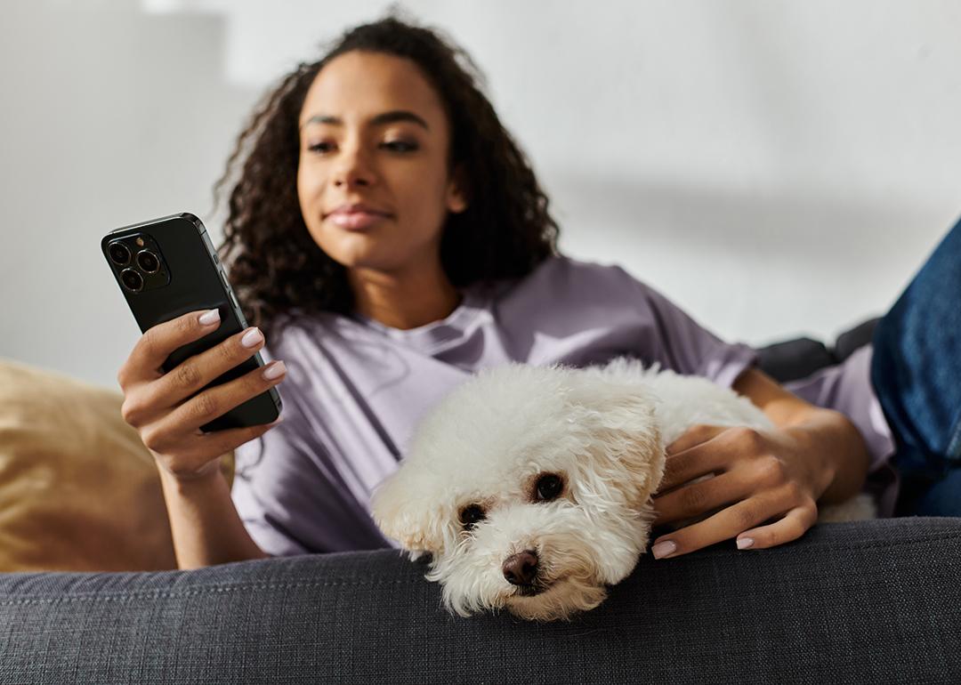 A woman relaxing on the couch while scrolling through her phone and petting her dog.