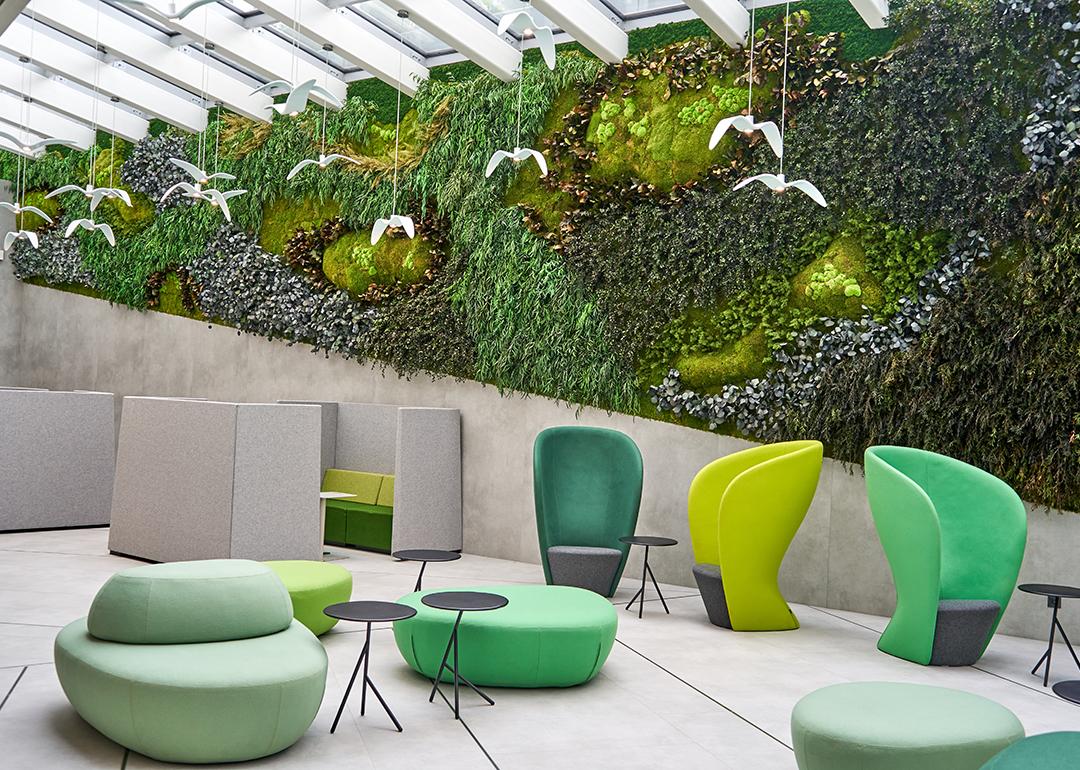 A modern office interior featuring a large biophilic living wall covered in various mosses, ferns, and plants spanning the back wall beneath a glass skylight ceiling with white beams. The space includes contemporary furniture in shades of green and lime.